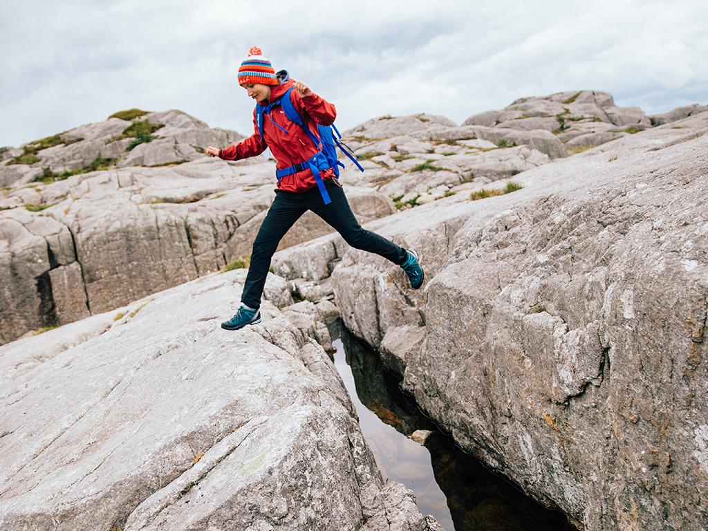 Kobieta w niskich butach trekkingowych skacząca przez skalną szczelinę wypełnioną wodą