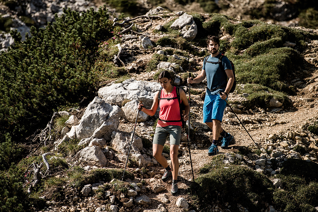 Dwoje turystów, którzy schodzą z gór. Mają na sobie koszulki, szorty i niskie buty trekkingowe, a w rękach kije trekkingowe.