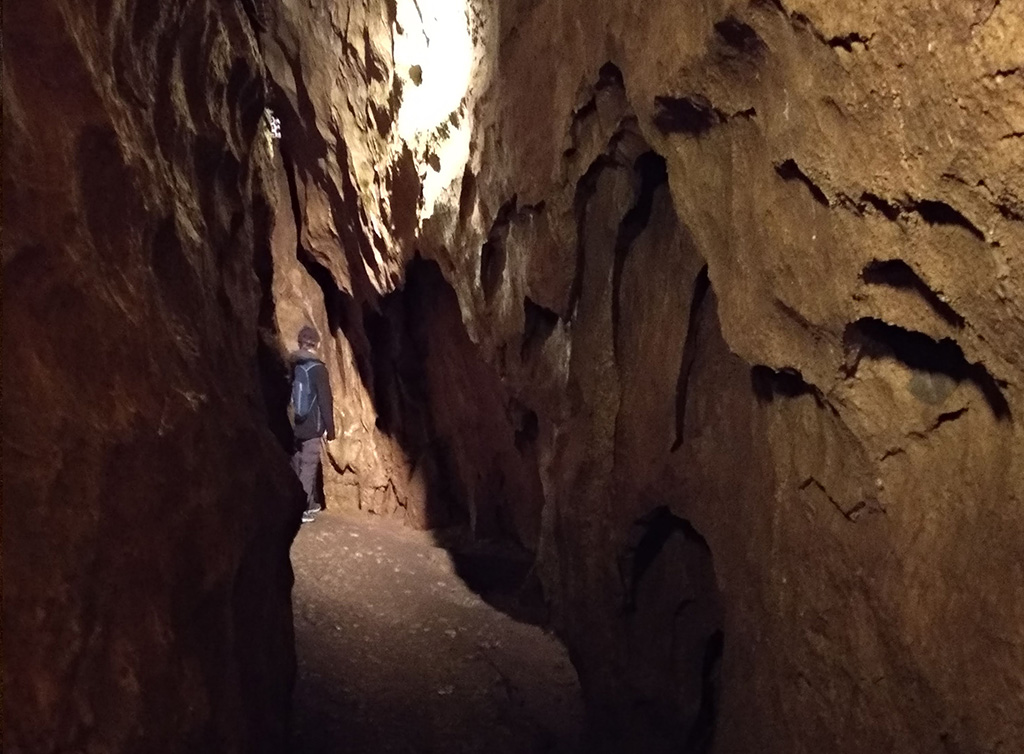 szczelina w jaskini na górze widać światło, na dalszym planie stoi chłopiec z plecakiem